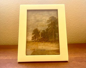 Op de afbeelding: Een wit fotolijstje met een landschapschilderij erin. Het schilderij toont een boslandschap met bomen en een rivier. Het lijstje is 5 inch bij 7 inch.