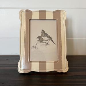 May include: A rectangular picture frame with a beige and brown striped border. The frame has a scalloped edge and holds a print of two birds perched on a decorative pot. The frame is made of a glossy material.