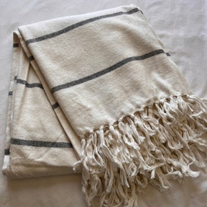 May include: A white throw blanket with grey stripes and a fringe border. The blanket is folded and lying on a white surface.