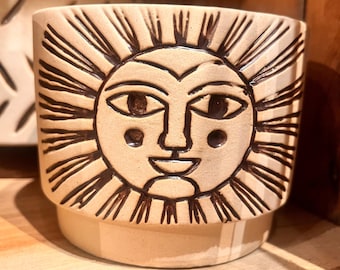 Beige colored ceramic planter with embossed sun face distressed rust color hold the 4 inch pot.