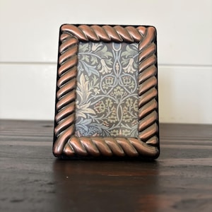 May include: A rectangular picture frame featuring a metallic copper-toned border with a twisted rope design. The frame surrounds a decorative insert printed with a repeating botanical pattern in shades of sage green, light blue, and cream.