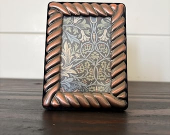 May include: A rectangular picture frame featuring a metallic copper-toned border with a twisted rope design. The frame surrounds a decorative insert printed with a repeating botanical pattern in shades of sage green, light blue, and cream.