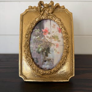 Puede incluir: Un marco de fotos dorado y ornamentado, con una abertura ovalada. El marco presenta un lazo en la parte superior y un diseño floral alrededor del óvalo. La imagen interior muestra un jarrón con flores rosas.