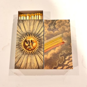 May include: Open matchbox with a vintage sun design on the front. The sun has a face and is surrounded by radiating lines. The box next to it has a rainbow and cloud design. The matches are visible inside the box.