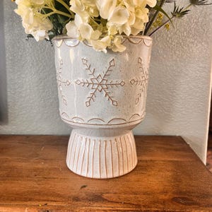 May include: A white ceramic planter with a snowflake design. The planter has a wide base and a narrow top. It is decorated with a raised snowflake pattern in a light brown colour.