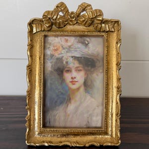 Puede incluir: Un marco dorado con un retrato de una mujer con un sombrero floral. El marco tiene un lazo decorativo en la parte superior y un diseño ornamentado. El retrato utiliza colores suaves y tiene un estilo vintage.