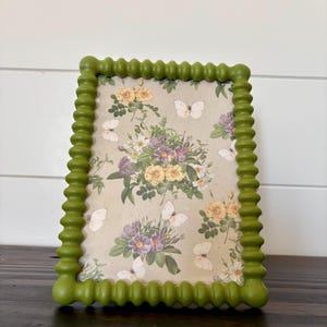 May include: A rectangular picture frame with a textured, beaded olive green border. The frame contains a print featuring a botanical illustration of yellow and purple flowers interspersed with white butterflies on a light beige background.