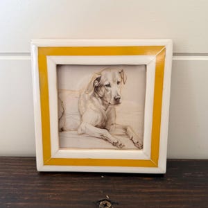 May include: A framed pencil sketch of a dog, featuring a light-colored Labrador Retriever lying down. The artwork is set within a white frame with a bright yellow border, creating a striking contrast. The frame is square and rests on a dark wooden surface.