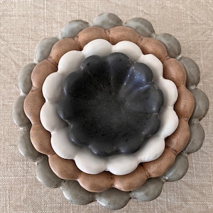 May include: A stack of ceramic dishes in a flower shape. The dishes are in shades of grey, brown, white, and black. The plates have a scalloped edge and are arranged in a nested design.