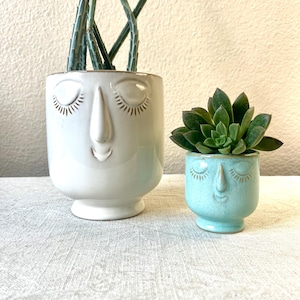 Może przedstawiać: Dwie ceramiczne donice z twarzami, jedna duża i biała, druga mała i turkusowa. Biała donica zawiera wysoką zieloną roślinę, a turkusowa - zielonego sukulenta. Obie donice mają rysy twarzy z zamkniętymi oczami.