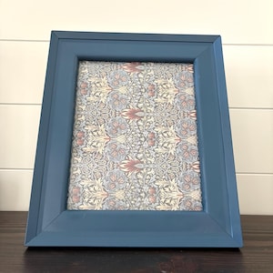 May include: A rectangular picture frame with a smooth, matte blue finish. The frame contains a decorative insert featuring a dense, repeating botanical pattern in shades of muted red, cream, and grey-blue, creating a detailed floral motif.
