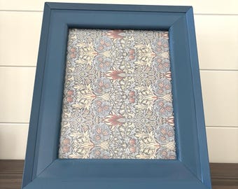 May include: A rectangular picture frame with a smooth, matte blue finish. The frame contains a decorative insert featuring a dense, repeating botanical pattern in shades of muted red, cream, and grey-blue, creating a detailed floral motif.