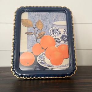 Puede incluir: Una impresión enmarcada que presenta una naturaleza muerta de naranjas, un jarrón con estampado azul y blanco y una ramita de hojas. El marco azul marino tiene un borde festoneado dorado. El fondo tiene un diseño floral azul y blanco.