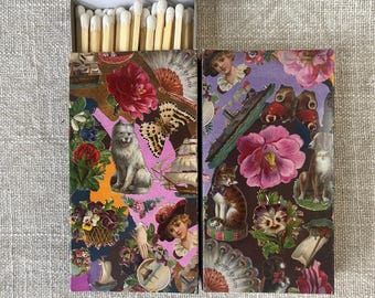 May include: Two decorative matchboxes with a collage design. One box features a gray dog, flowers, and a butterfly. The other box has a cat, flowers, and a dog. The match heads are white.