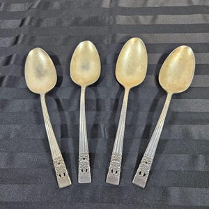 May include: Four antique silver spoons with oval bowls and detailed handles. The handles have a ribbed design with floral accents near the base. The spoons are arranged on a black striped surface.