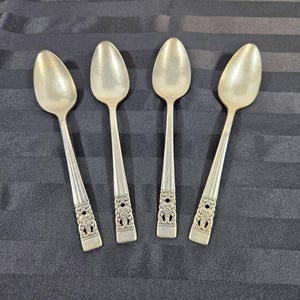 May include: Four silver spoons with oval bowls and ornate floral designs on the handles. The spoons are arranged on a dark, textured surface, showcasing their reflective quality and intricate details.