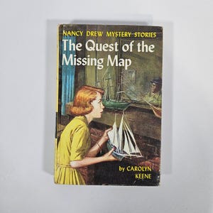 May include: A vintage hardcover book titled "The Quest of the Missing Map" by Carolyn Keene. The cover features an illustration of a young woman in a yellow dress looking at a model sailboat. The book is part of the Nancy Drew Mystery Stories series.