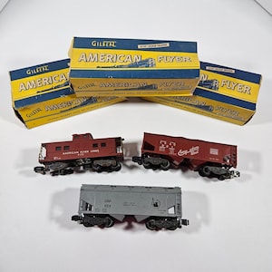 May include: A collection of vintage Gilbert American Flyer model train cars and boxes. The boxes are yellow and blue with the words "American Flyer" printed on them. The train cars are red and gray.