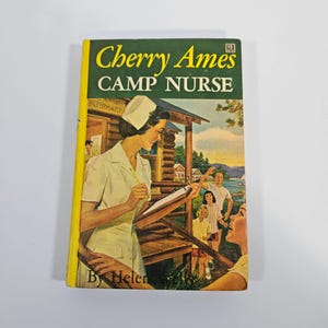 May include: Vintage paperback book titled "Cherry Ames Camp Nurse" by Helen Wells. The cover features an illustration of a nurse in a white uniform and cap, holding a clipboard, with a camp scene in the background. The book has a yellow spine.