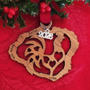 May include: A wooden Christmas ornament in the shape of the Kauai island, with heart cutouts and the year 2026. The ornament is made of koa wood and has the words "KAUAI" and "WOOD" on it. It hangs from a red ribbon.