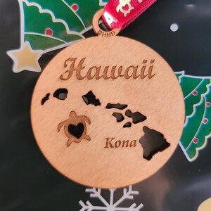 May include: A round wooden ornament with the word "Hawaii" in a script font. The ornament features cutouts of the Hawaiian islands and a turtle with a heart-shaped cutout. The word "Kona" is below the islands. A red ribbon is attached for hanging.