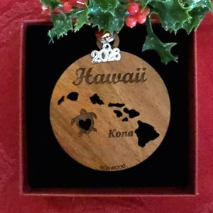 May include: Wooden Hawaii ornament with a map outline, a turtle with a heart, and the word "Kona." The year "2023" is displayed above. The ornament is brown and is displayed in a red box with holly.