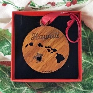 May include: A wooden ornament shaped like the islands of Hawaii with a turtle and a heart cutout. The ornament is in a red box with a black velvet lining. The ornament is made of Koa wood and says "Hawaii" and "Koa Wood" on it.