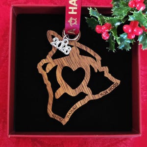 May include: A wooden Hawaii ornament in a gift box. The ornament is shaped like the island of Hawaii with a heart cutout. It has the text "HAWAII" and "KOA WOOD" on it, and a small charm that says "2026". A red ribbon is attached.