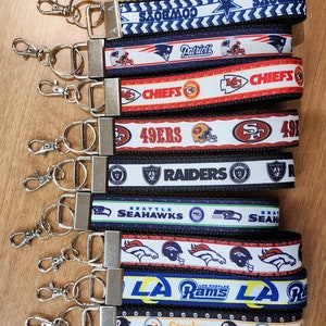 May include: Ten keychains with different NFL team logos. The keychains are black and white with red, blue, and yellow accents. The logos include the Cowboys, Patriots, Chiefs, 49ers, Raiders, Seahawks, Broncos, Rams, and Steelers.