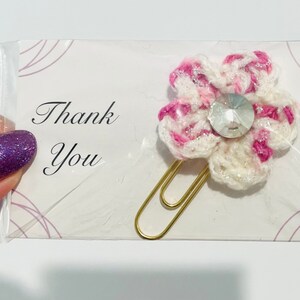 May include: A handmade flower-shaped paper clip with a crocheted design in white, pink, and silver. The flower has a clear gem center and is attached to a gold-colored paper clip. The card reads "Thank You".