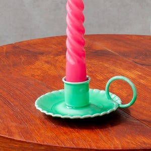 Könnte beinhalten: Eine brennende, rosa, spiralförmige Kerze in einem grünen Metallkerzenhalter mit Griff. Der Kerzenhalter steht auf einem runden Holztisch. Rechts von der Kerze befindet sich eine Glasvase mit Wasser und Grün.