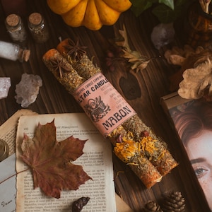 Può includere: Una composizione mostra una piccola zucca arancione, foglie secche e due candele da altare. Le candele sono avvolte in un'etichetta rosa con testo. Sono presenti anche piccoli barattoli di vetro e un libro vintage, creando un tema autunnale.
