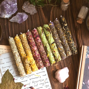 Op de afbeelding: Een assortiment kleurrijke kaarsen, versierd met gedroogde kruiden en bloemen, gerangschikt op een houten oppervlak. De kaarsen worden vergezeld door kristallen, vintage papier en een decoratieve doos met een heksenillustratie. Een messing kom en glazen potten zijn ook aanwezig.