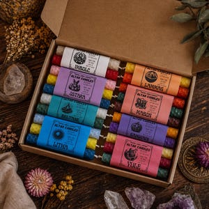 Könnte beinhalten: Eine Schachtel mit Altar-Kerzen mit farbigen Etiketten. Jedes Etikett trägt einen anderen Namen: Imbolc, Ostara, Beltane, Litha, Lammas, Mabon, Samhain und Yule. Die Kerzen sind in verschiedenen Farben, darunter Rot, Gelb, Blau und Grün.