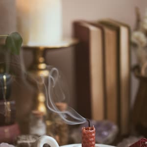 May include: A red candle, recently extinguished, sits in a white ceramic holder. Wisps of smoke curl upwards. The background includes a large lit candle, books, crystals, and small glass bottles, suggesting a spiritual or decorative theme.