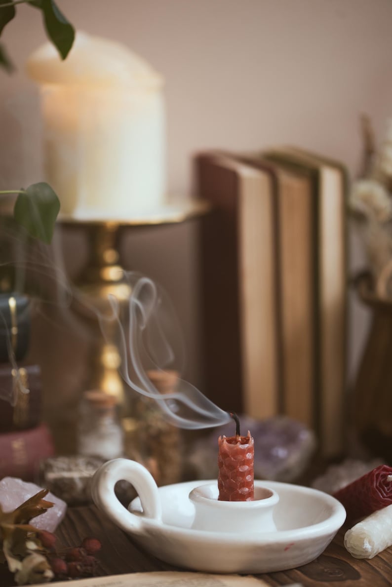 May include: A red candle, recently extinguished, sits in a white ceramic holder. Wisps of smoke curl upwards. The background includes a large lit candle, books, crystals, and small glass bottles, suggesting a spiritual or decorative theme.