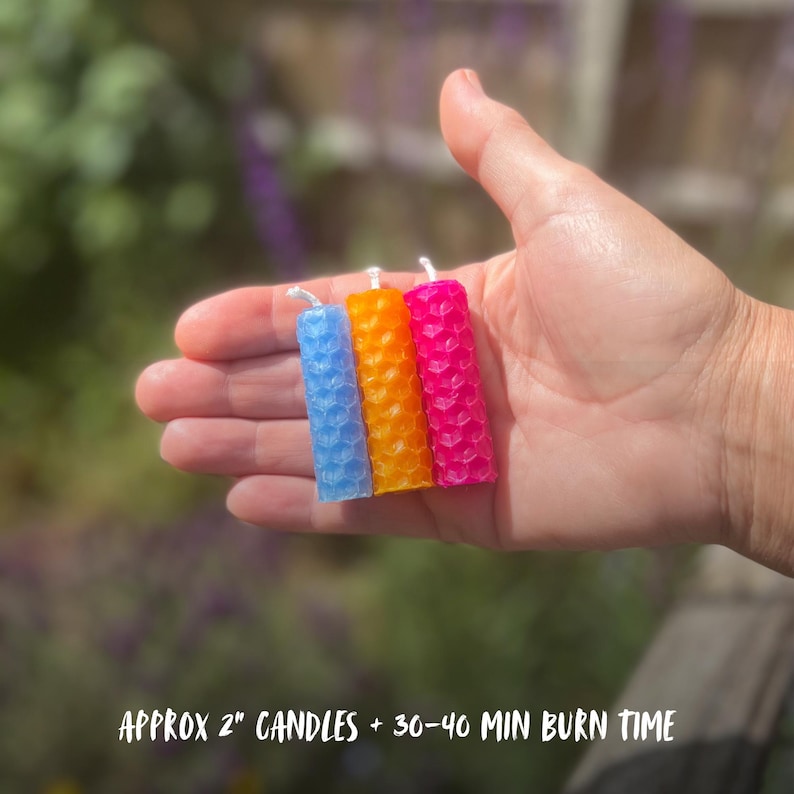 May include: Three small, colorful candles in blue, orange, and pink, held in a hand. The candles are approximately 2 inches tall and have a honeycomb texture. Each candle has a white wick. The background is blurred, with green and purple hues.