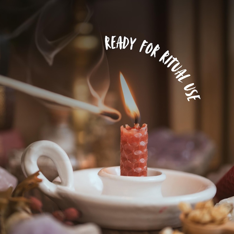 May include: A lit red candle in a white ceramic holder. The candle has a textured surface and is topped with a flame. The text "READY FOR RITUAL USE" is arched above the candle. A lit match is held near the candle.