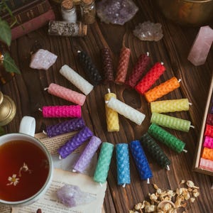 May include: A collection of colorful, cylindrical beeswax candles arranged on a wooden surface. The candles are in various colors, including pink, blue, green, and yellow. Other items include crystals, a cup of tea, and dried flowers.