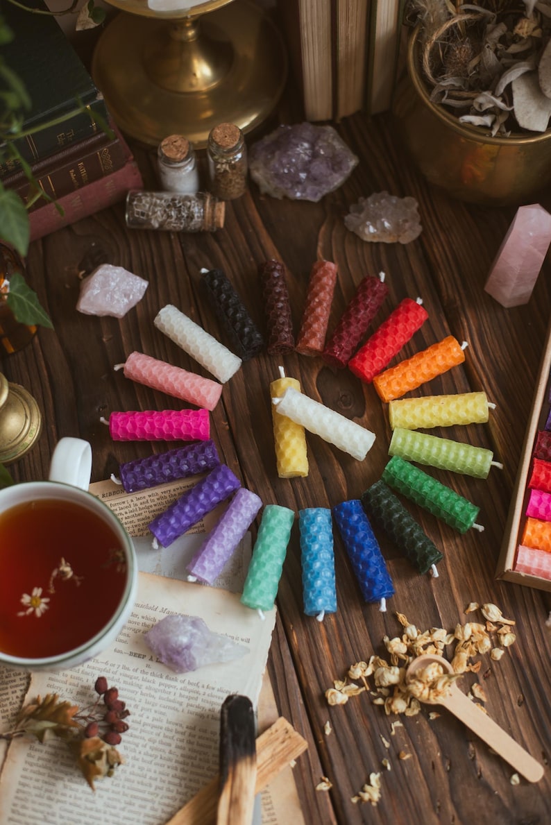 May include: A collection of colorful, cylindrical beeswax candles arranged on a wooden surface. The candles are in various colors, including pink, blue, green, and yellow. Other items include crystals, a cup of tea, and dried flowers.