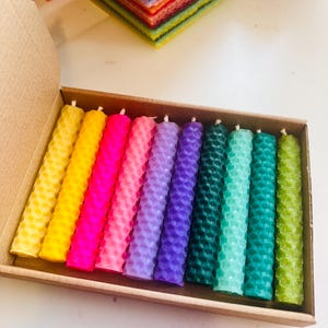 Könnte beinhalten: Ein Set mit 12 bunten Honigwaben-Bienenwachskerzen in einer Kartonbox. Die Kerzen sind in einem Regenbogenmuster angeordnet, wobei jede Kerze etwa 10 cm lang ist.
