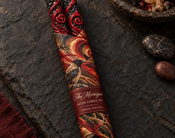 May include: Two black honeycomb-textured candles with red wax swirls, wrapped in a paper sleeve featuring a crow illustration and red swirling patterns. The label reads The Morrigan Altar Candle Set Phoenix Heritage in white serif lettering.
