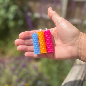 An outdoor shot of a woman's open hand holding three beeswax spell candles (light blue, orange and pink) to show the size. Candles sit vertically in the palm and stretch from the edge of the index finger down to the little finger.