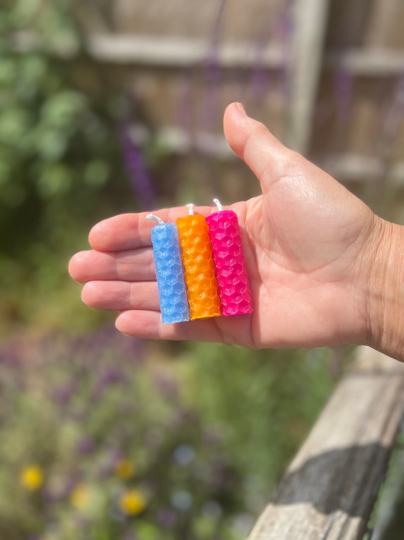 An outdoor shot of a woman's open hand holding three beeswax spell candles (light blue, orange and pink) to show the size. Candles sit vertically in the palm and stretch from the edge of the index finger down to the little finger.