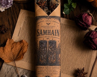 May include: Two black pillar candles wrapped in brown paper packaging labeled Phoenix Candle Company Samhain. The candles are topped with dried herbs, star anise, and small amethyst crystals. The label lists ingredients including thistle, orange peel, star anise, and amethyst, noting hand-rolled beeswax and a cotton wick.