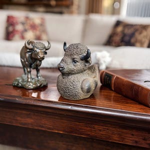 Two bison figurines on a wooden table. One is a bronze-colored standing sculpture, and the other is a brown, textured duck-shaped figurine. A leather-bound book rests nearby. The background includes a white couch and decorative pillows.