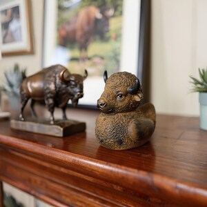 A brown bison-shaped resin duck with black horns and a detailed textured surface. A bronze bison statue is in the background. The duck is on a wooden surface.
