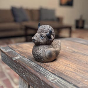 May include: A brown rubber duck shaped like a bison. The duck has a textured body, small black horns, and a detailed face. It is sitting on a wooden table.