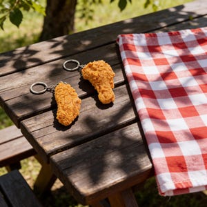 Pair of realistic fried chicken wing keychains with silver key rings displayed on a wooden picnic table with a red-and-white gingham cloth and warm sunlight—cute novelty food keychains for wing fans, bag charms, and quirky gift ideas.