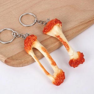 May include: Two keychains shaped like chicken drumsticks with a red and orange glaze. The keychains are made of plastic and have a realistic look.